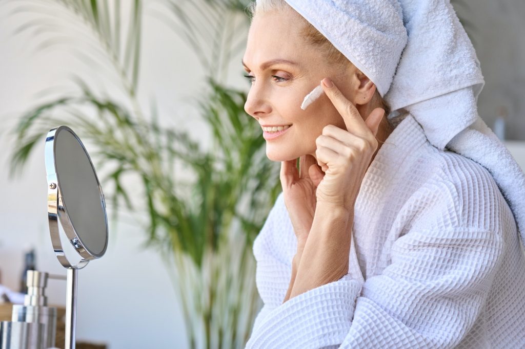 Smiling beautiful older lady looking at mirror applying cream. Skincare concept.