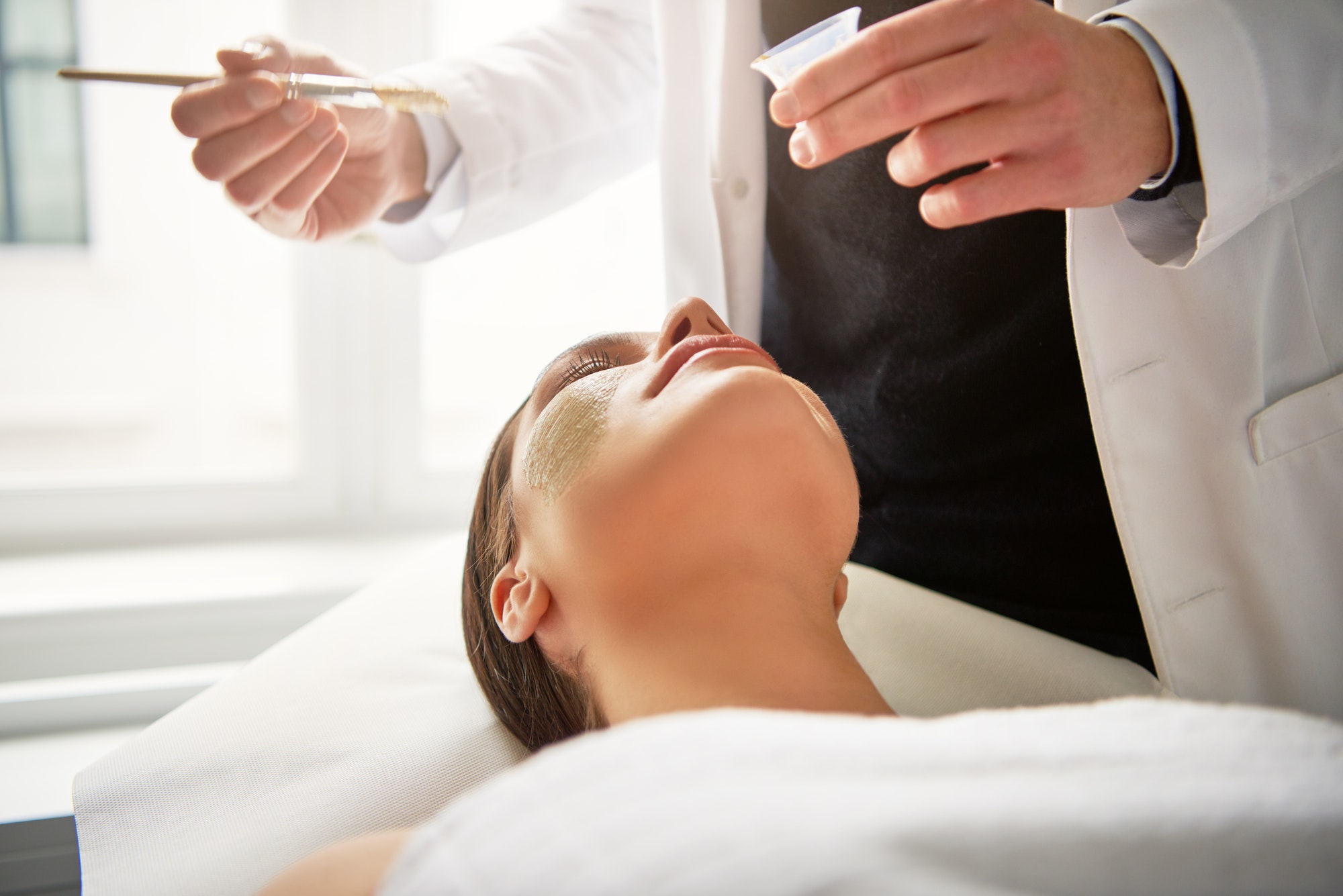 Beautician putting mask on young female face in salon