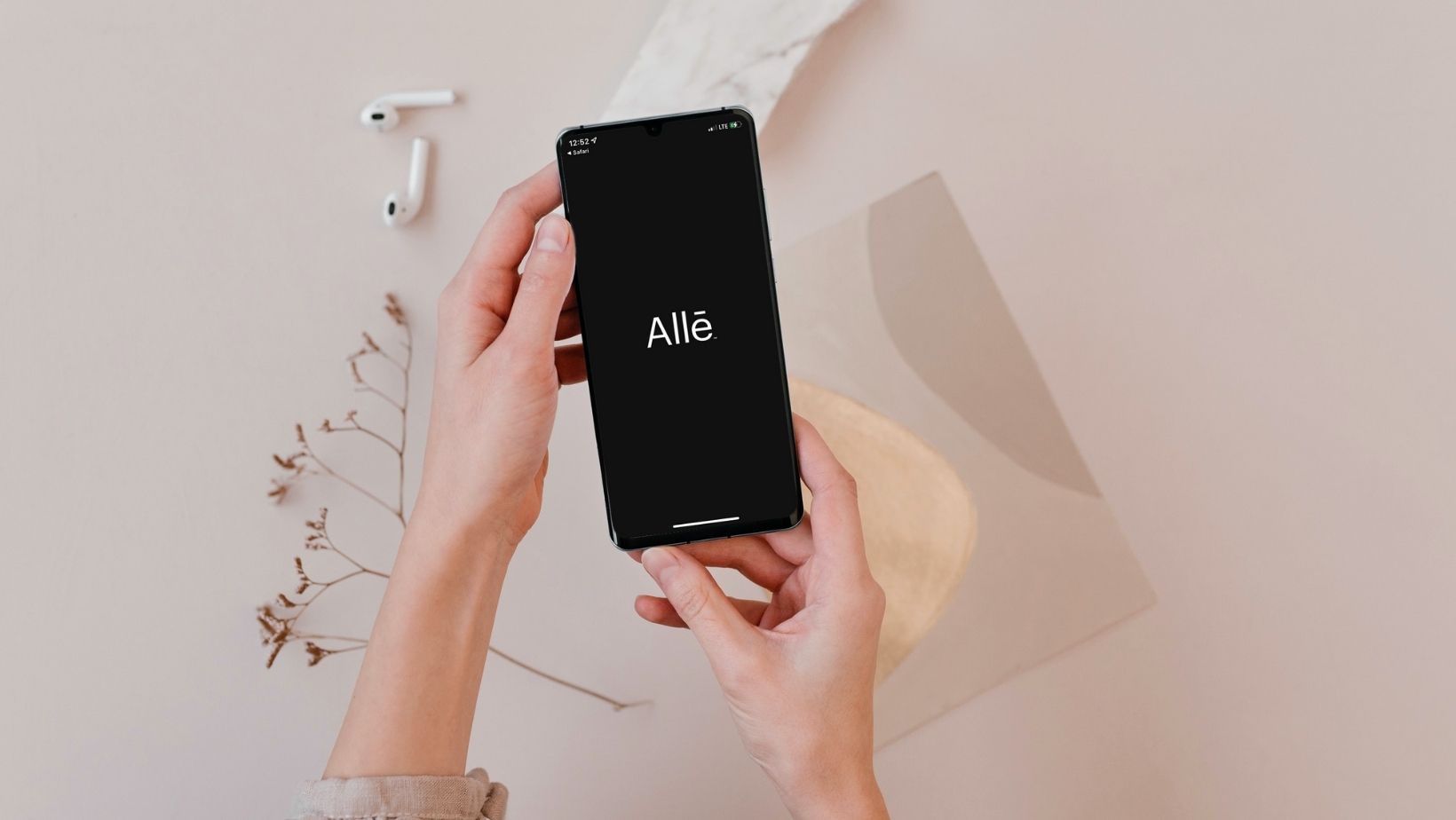 Hands holding a smartphone with the Allē app logo on the screen, ready for use in a minimal and modern setting with AirPods and neutral decor.