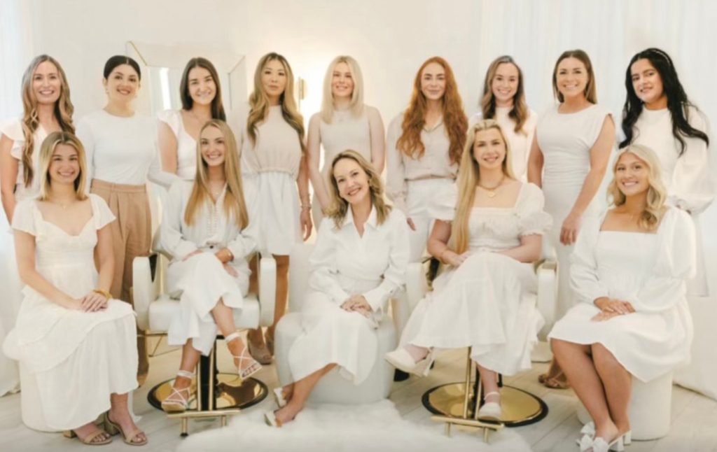 Group of women dressed in white and light neutral tones, seated and standing in a bright, elegant space with soft lighting.
