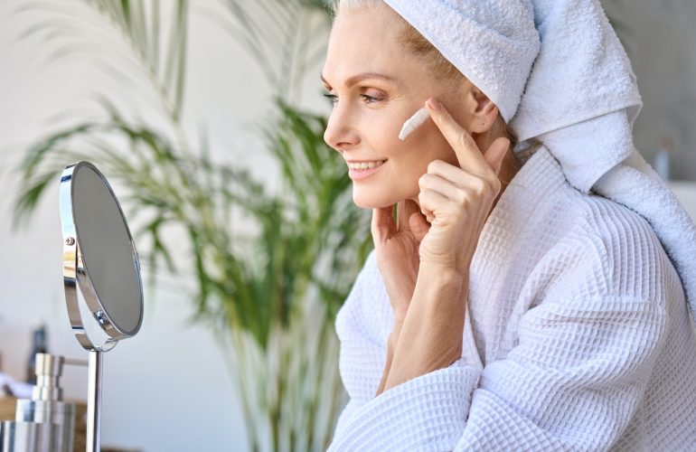 Smiling beautiful older lady looking at mirror applying cream. Skincare concept.